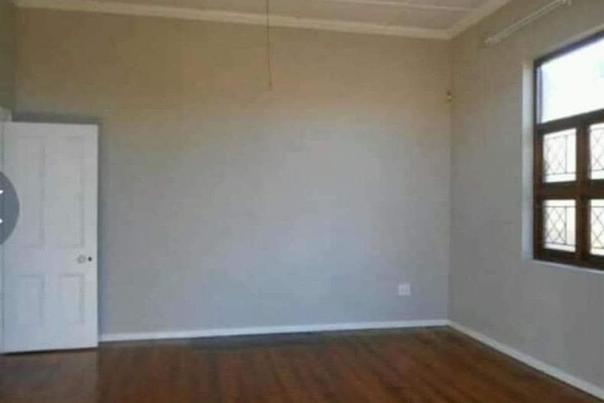 A spacious, empty living room featuring timber flooring, a ceiling fan, and a window with security bars.