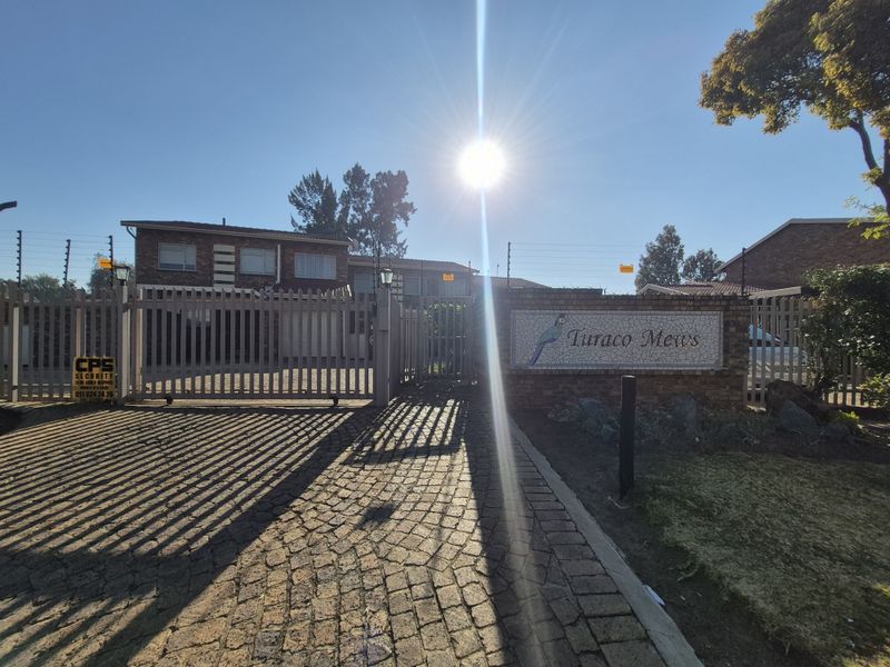 Front entrance view of Turaco Mews townhouse complex featuring a paved driveway, secure sliding gate, and brick boundary wall with electric fencing.