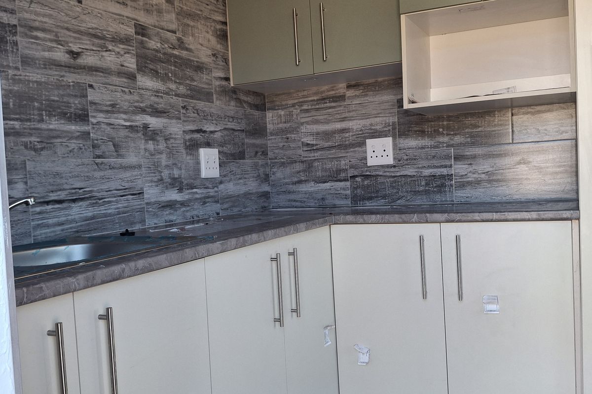A modern, compact kitchen featuring light-toned cabinetry, wood-look tiled backsplash, and a stainless steel sink with clean, contemporary finishes.
