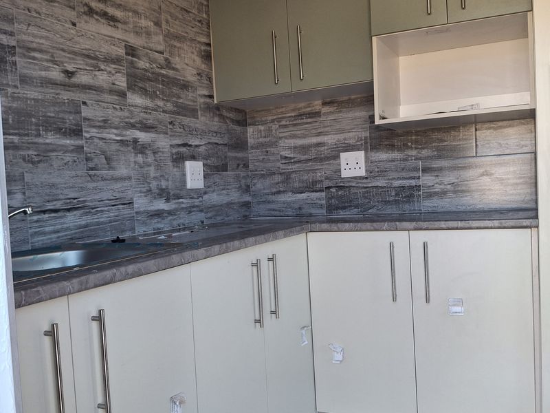 A modern, compact kitchen featuring light-toned cabinetry, wood-look tiled backsplash, and a stainless steel sink with clean, contemporary finishes.