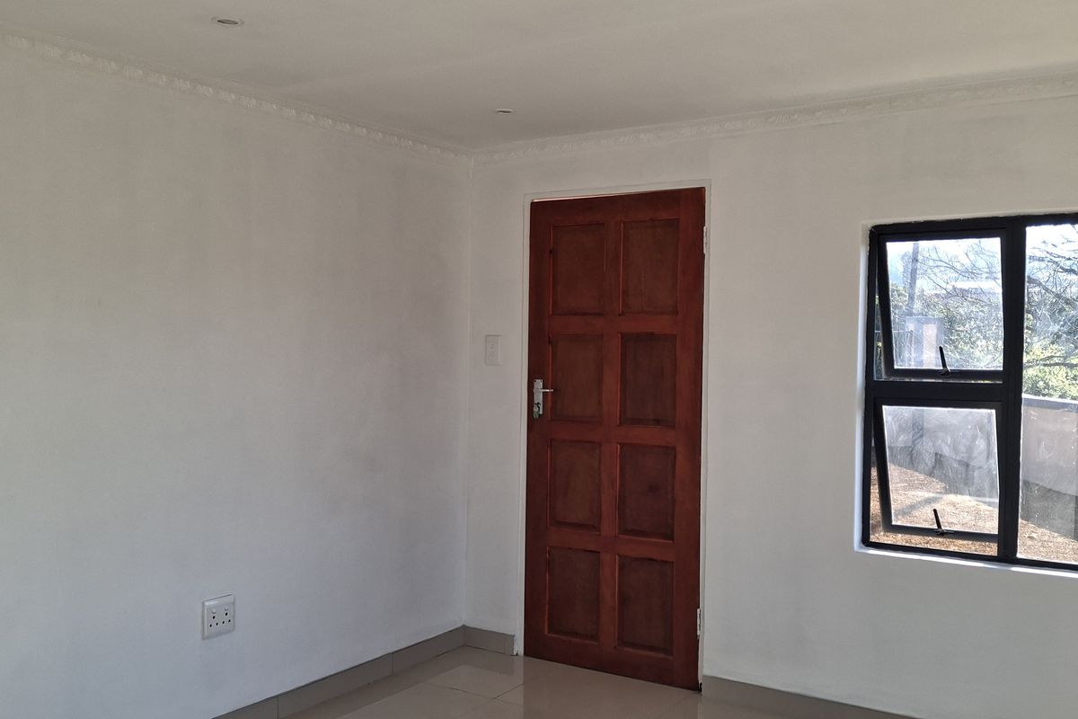A clean, empty bedroom featuring neutral tiled flooring, a solid wooden door, and an aluminium-framed window with natural light.