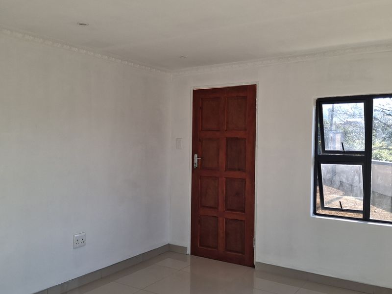A clean, empty bedroom featuring neutral tiled flooring, a solid wooden door, and an aluminium-framed window with natural light.