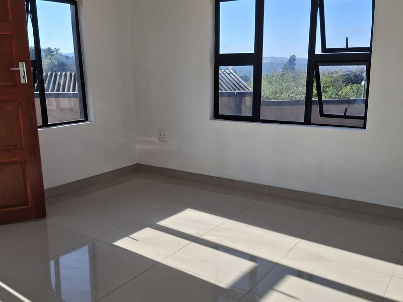 A bright, empty bedroom featuring neutral tiled flooring, modern dark aluminium window frames, and a solid wooden door.