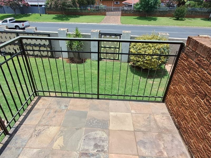 An elevated balcony featuring stone tile flooring, a metal balustrade, and a brick wall, overlooking the front garden and street.