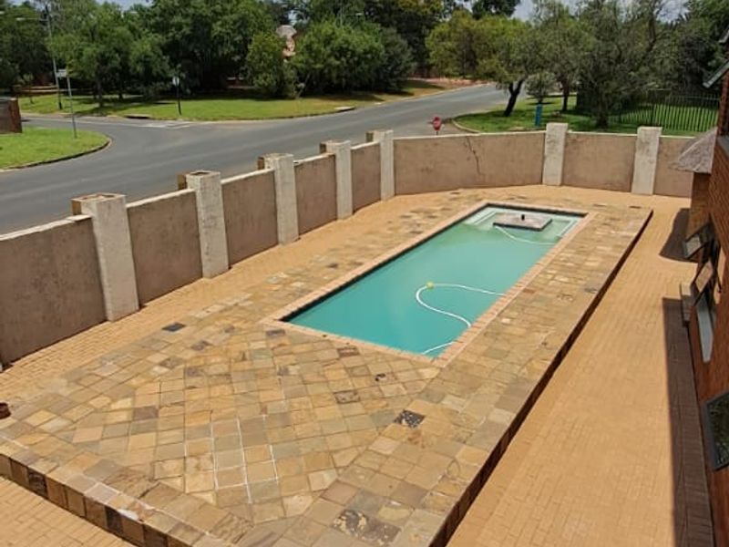 Outdoor pool area featuring a rectangular swimming pool surrounded by stone and brick paving, enclosed by a perimeter boundary wall.