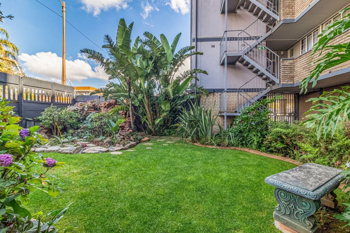 A well-maintained rear garden area featuring a lush lawn, established rockery, and direct access to the apartment building via a metal fire escape staircase.