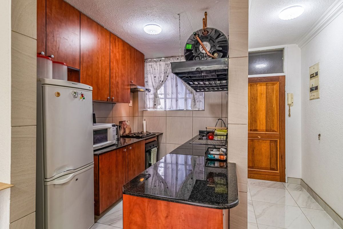 Compact kitchen featuring dark granite countertops, wooden cabinetry, built-in oven, hob, and tiled flooring, adjacent to the apartment entrance.