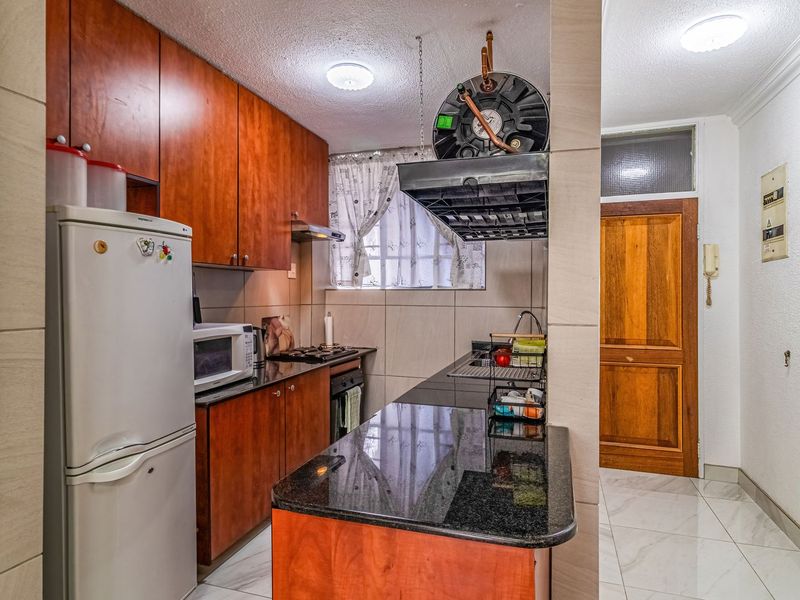 Compact kitchen featuring dark granite countertops, wooden cabinetry, built-in oven, hob, and tiled flooring, adjacent to the apartment entrance.