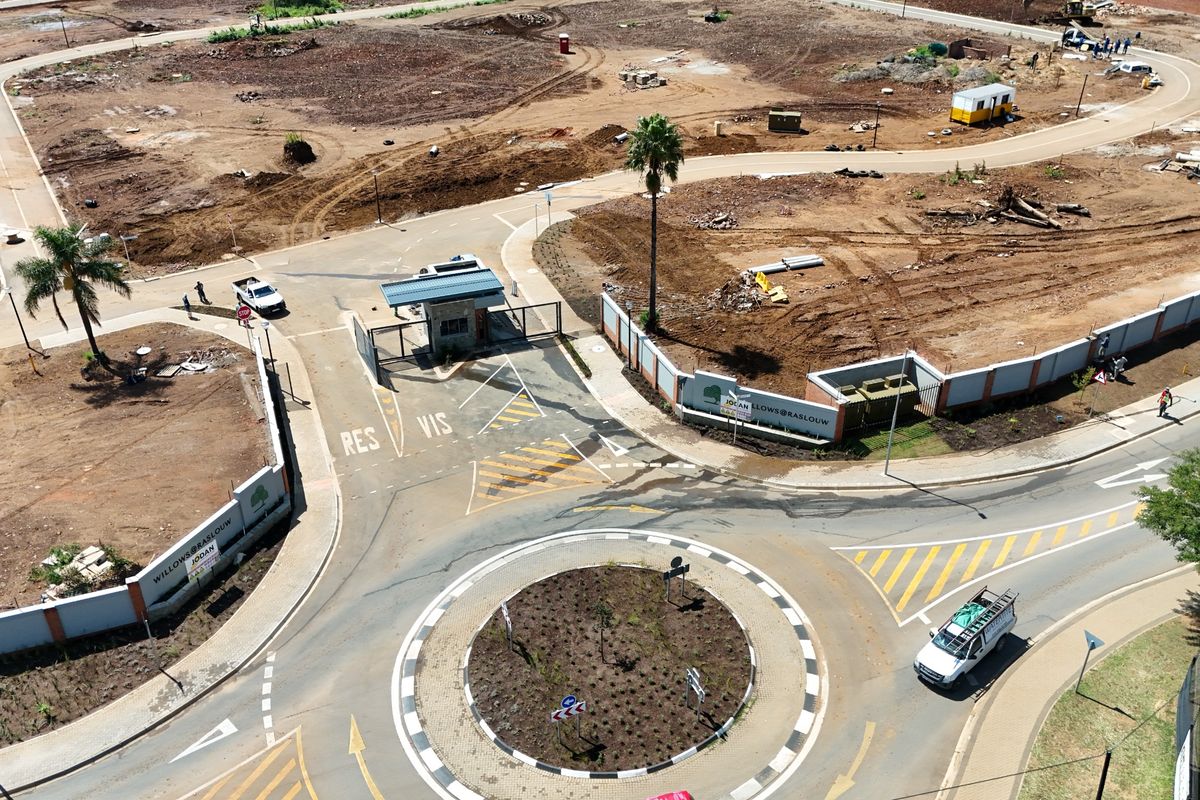 Aerial view of a residential estate entrance featuring a security gatehouse, perimeter walling, and a paved roundabout access point.