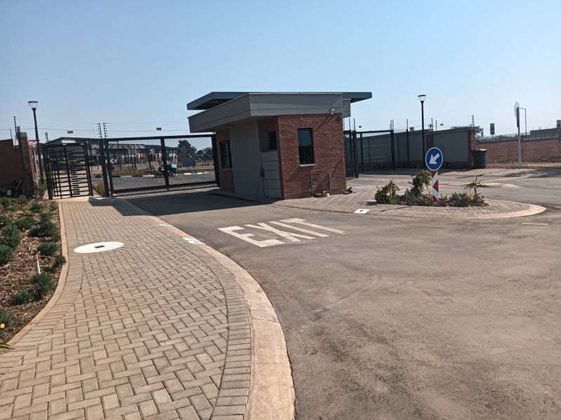 A secure residential estate entrance featuring a brick guard house, automated security gates, paved pedestrian walkways, and asphalt driveway access.