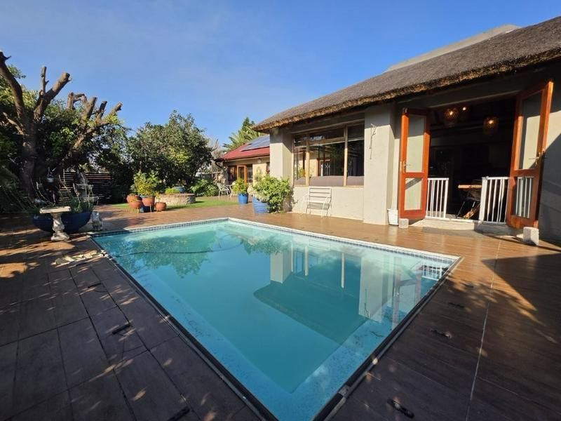 A well-maintained outdoor pool area featuring a tiled deck, thatched roof structure, and direct access to the home through double doors.