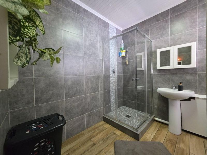 A modern bathroom featuring grey wall tiles, a glass shower enclosure, a pedestal basin, and wood-look floor tiles.
