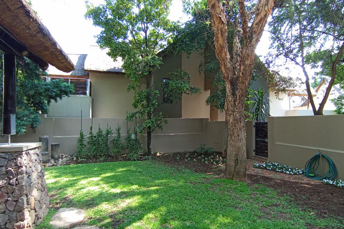 A private rear garden featuring a well-maintained lawn, mature trees, and a stone-clad pillar under a thatched roof structure.