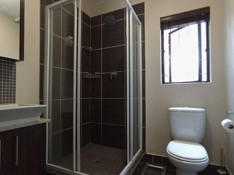 A functional bathroom featuring a shower enclosure, vanity basin, and toilet with dark tiled accents and a window for natural light.