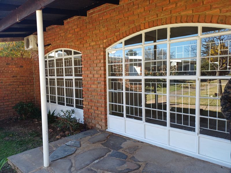 A covered patio area featuring flagstone flooring, exposed brick walls, and large arched steel-framed windows with integrated security bars.