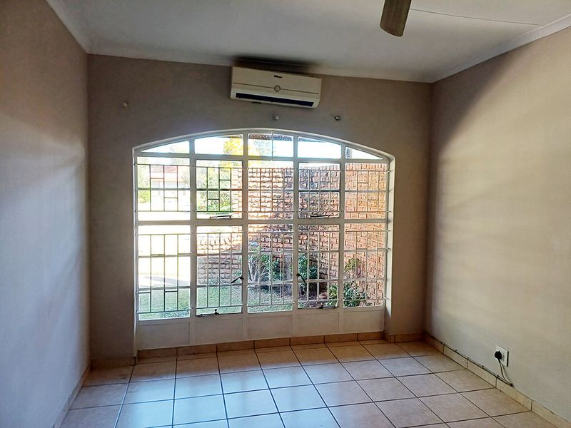 An empty living room featuring tiled flooring, a wall-mounted air conditioning unit, a ceiling fan, and a large arched window with security bars.