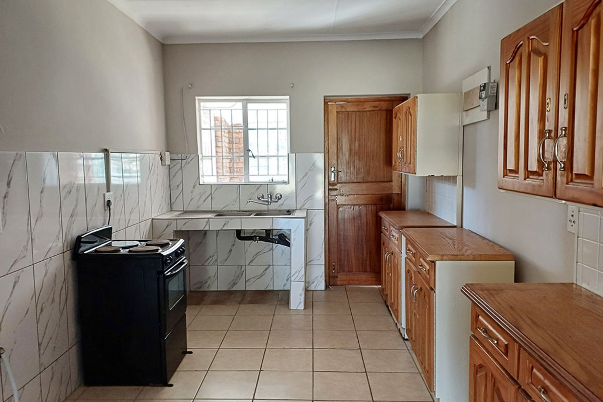 A functional kitchen featuring tiled splashbacks, wooden cabinetry, tiled flooring, and a window with security bars, offering a clean and practical layout.
