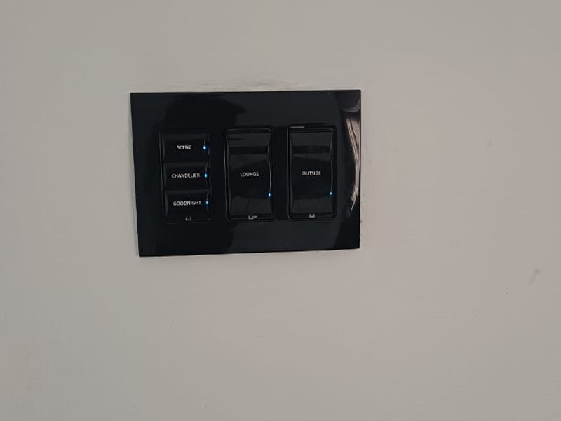 A modern black smart lighting control panel mounted on a white interior wall, featuring multiple programmable scene settings.