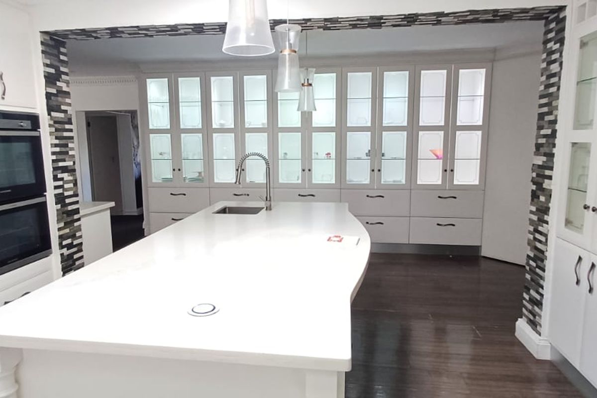 Modern kitchen featuring a large central island, built-in double oven, extensive glass-front cabinetry with integrated lighting, and dark hardwood flooring.