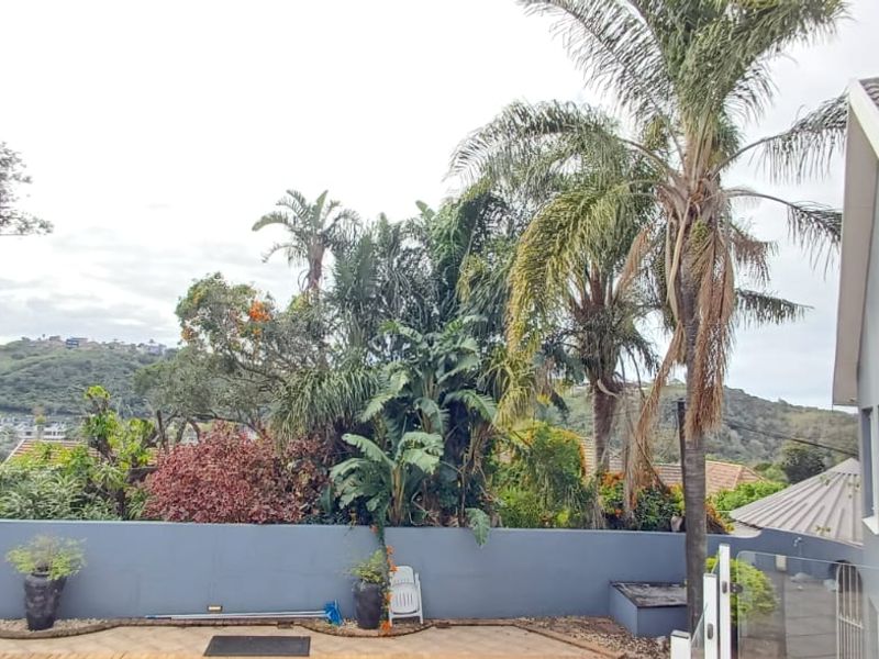 Rear exterior view featuring a paved patio area, glass balustrade, and a retaining wall overlooking a lush garden and hillside landscape.
