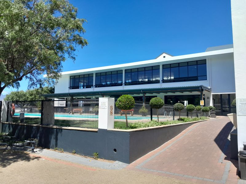 Exterior view of a communal swimming pool area with security fencing, paved access path, and large windows overlooking the facility.