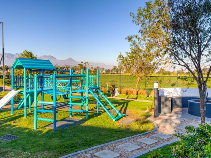 A well-maintained communal garden area featuring a children's playground, built-in circular seating, and paved pathways with mountain views.