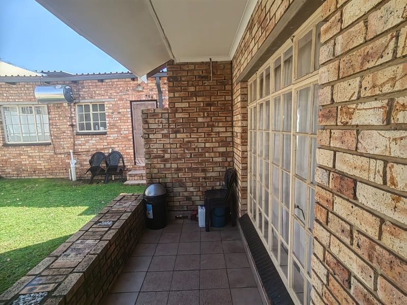 A covered brick patio area featuring tiled flooring and large multi-pane windows, overlooking a small private garden space.
