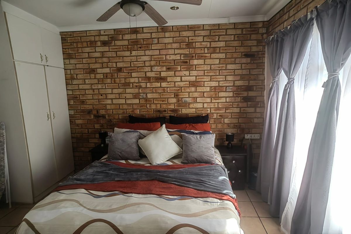A bedroom featuring an exposed brick accent wall, built-in cupboards, tiled flooring, and a ceiling fan, offering a warm and textured aesthetic.