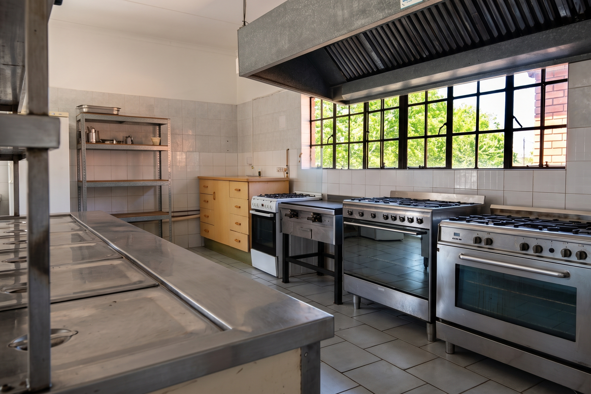 A functional commercial-style kitchen featuring multiple stainless steel gas stoves, a large extractor hood, tiled surfaces, and ample stainless steel preparation counters.