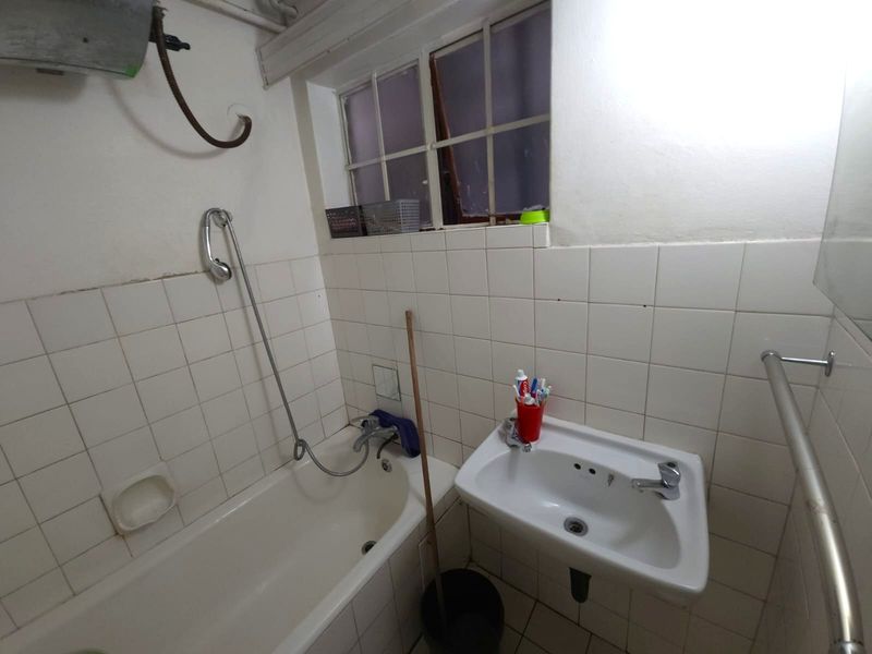A compact bathroom featuring a bathtub with shower mixer, wall-mounted basin, and tiled walls, requiring cosmetic updates to improve its overall condition.
