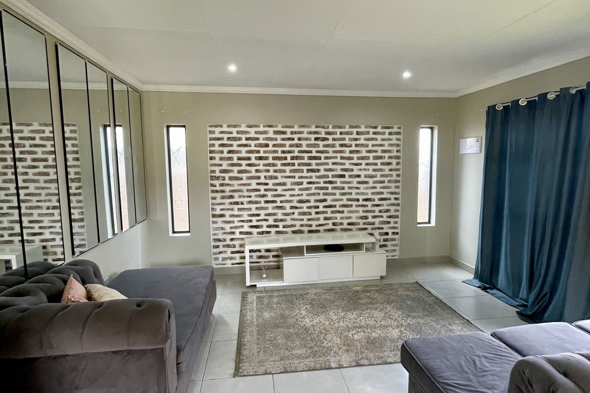 A well-lit living room featuring a distinctive exposed brick accent wall, mirrored wall panels, and neutral tiled flooring throughout.