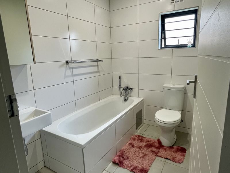 A clean, modern bathroom featuring white wall tiles, a built-in bathtub, a wall-mounted basin, and a standard toilet.