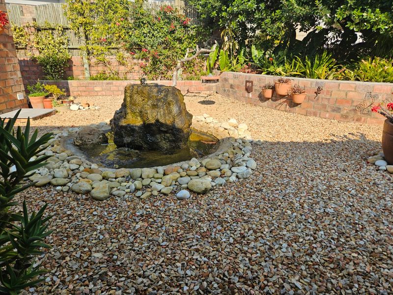 A landscaped garden area featuring a central stone water feature, gravel ground cover, and a brick retaining wall with established greenery.