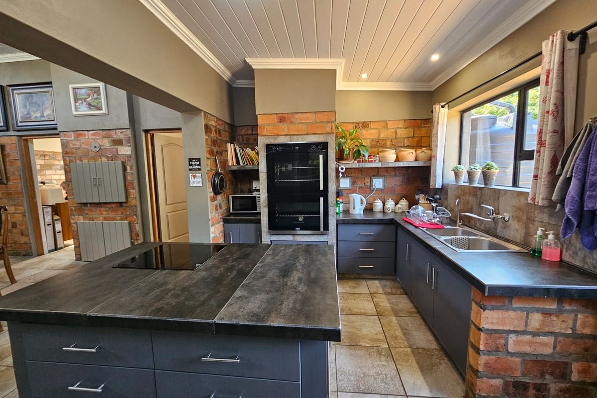 A well-appointed kitchen featuring a central island with integrated hob, built-in double oven, dark cabinetry, and rustic exposed brick accents.