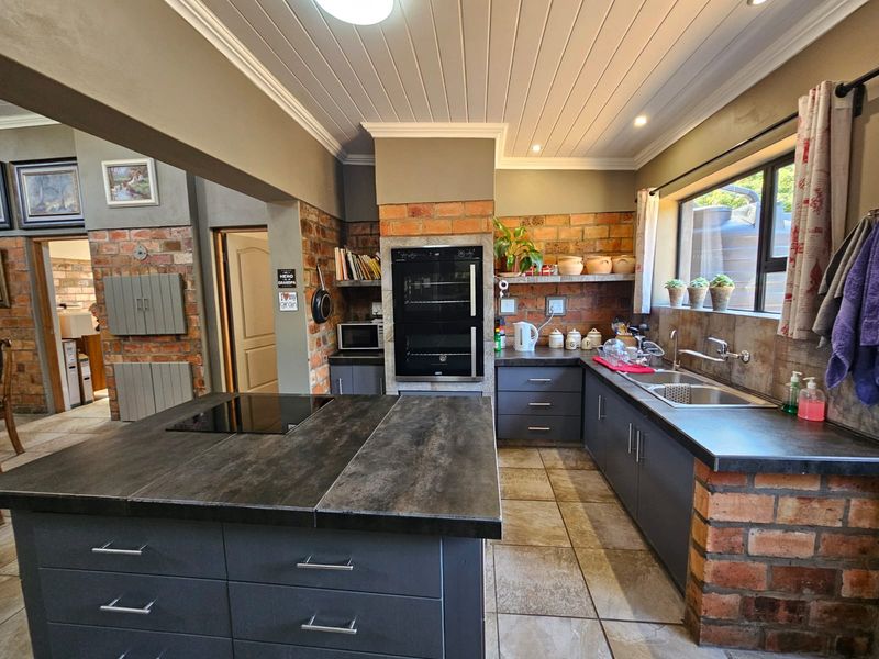 A well-appointed kitchen featuring a central island with integrated hob, built-in double oven, dark cabinetry, and rustic exposed brick accents.