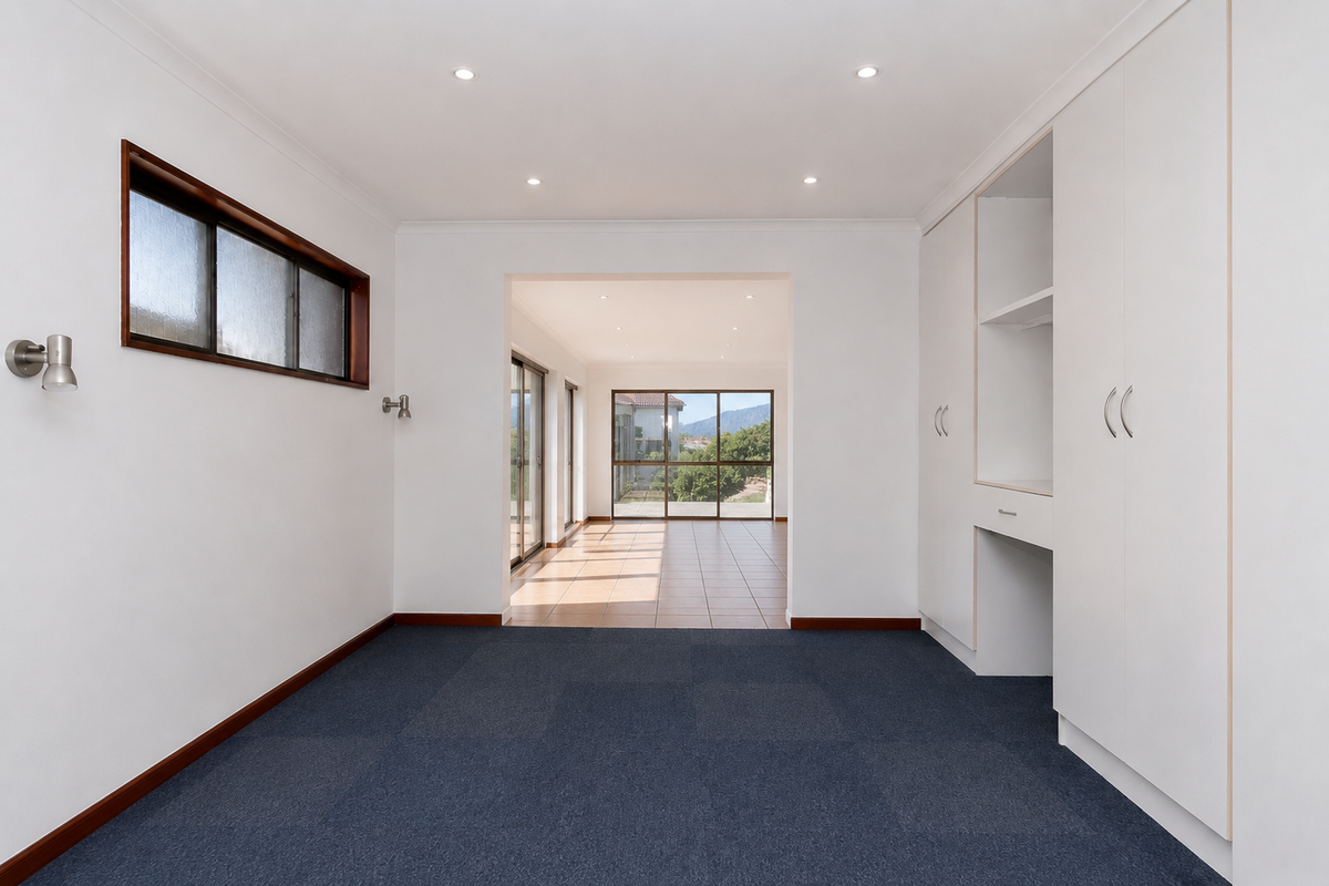 A bright, empty bedroom featuring built-in cupboards, recessed lighting, and carpet flooring, with a clear view through to an adjacent sunroom.