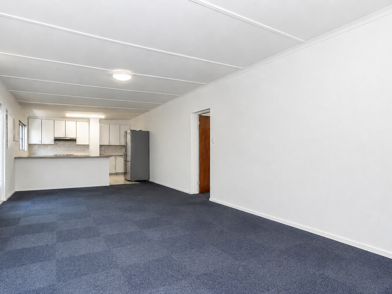 An open-plan living and kitchen area featuring neutral walls, blue carpet flooring, and a functional kitchen with built-in cabinetry and a breakfast bar.