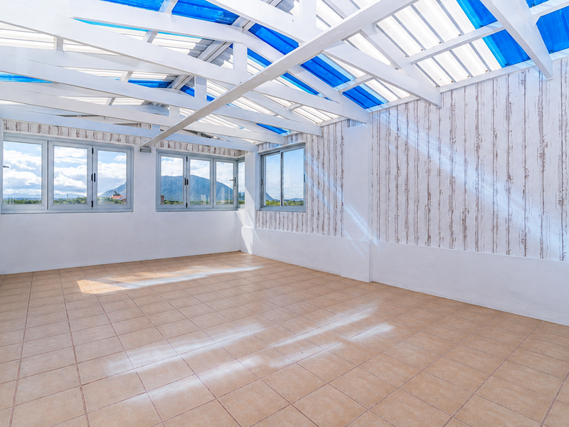 An empty, sunlit enclosed patio or sunroom featuring exposed timber roof trusses, translucent blue roof panels, and tiled flooring with mountain views.