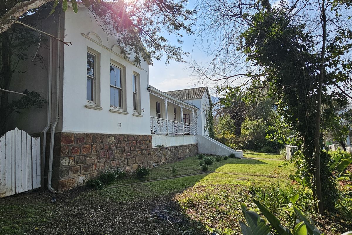 A side view of a historic-style house featuring stone cladding, a covered veranda with wrought iron railings, and mature garden surroundings.