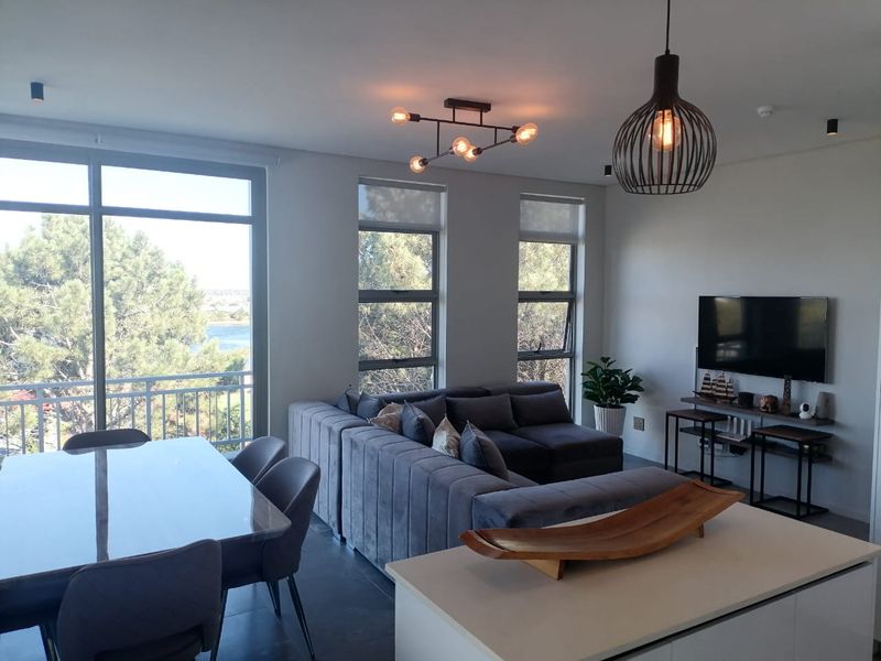 A bright and modern open-plan living and dining area featuring large windows, contemporary lighting, and access to a balcony with scenic views.