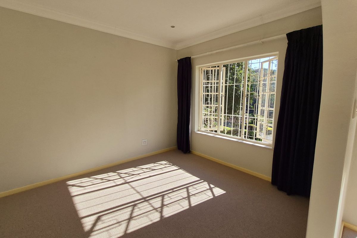 A bright, empty bedroom featuring neutral carpeting, cornice detailing, and large windows fitted with security bars and dark curtains.