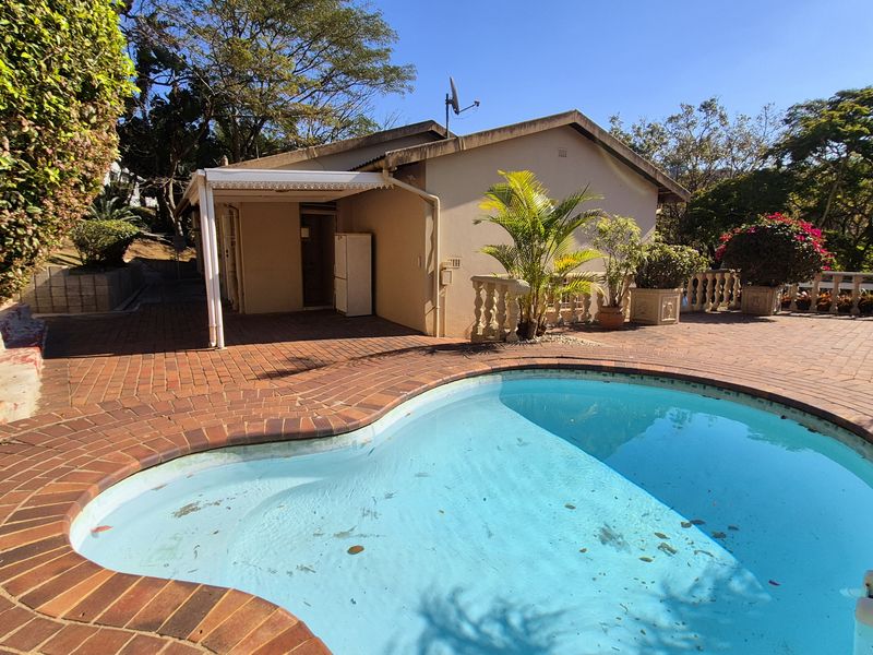 Outdoor pool area featuring a swimming pool, brick paved surround, and a covered patio area, set within a landscaped garden environment.