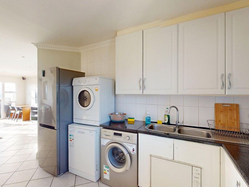 A functional kitchen area featuring white cabinetry, granite countertops, a double basin sink, and tiled finishes, seamlessly flowing into the adjacent living space.