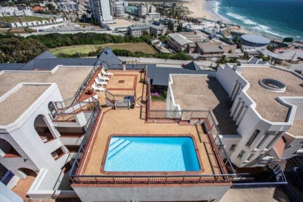 Aerial view of an apartment complex rooftop featuring a swimming pool, paved terrace area, and metal safety railings with coastal views.