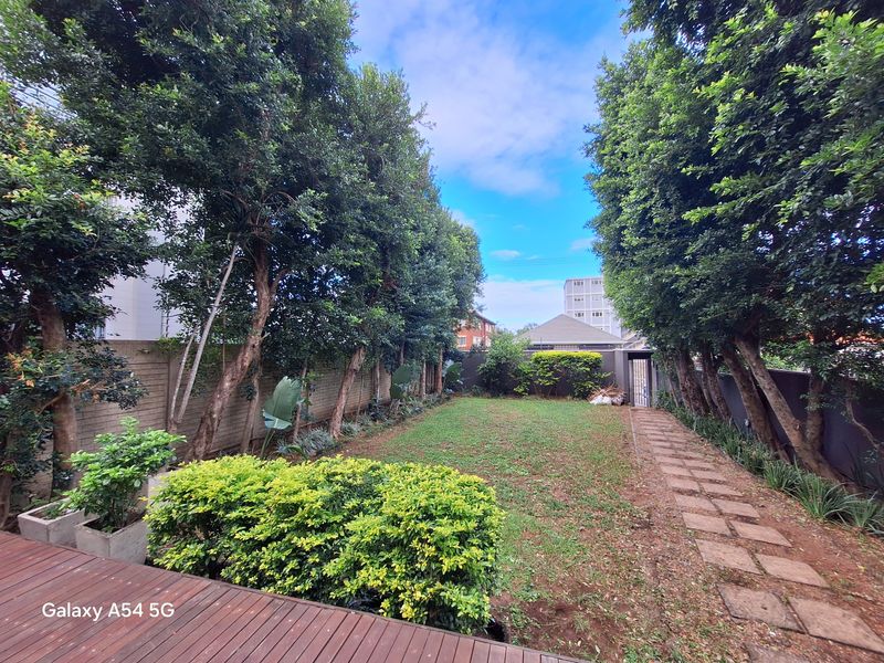 A private garden area featuring a timber deck, stone paver pathway, and established perimeter trees with a lawn space.