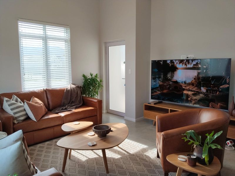 A bright and modern living room featuring tiled flooring, a glass panel door, and large windows fitted with venetian blinds.