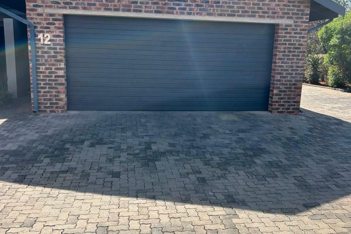 A well-maintained townhouse garage featuring a dark sectional door, exposed brick facade, and a paved driveway area.