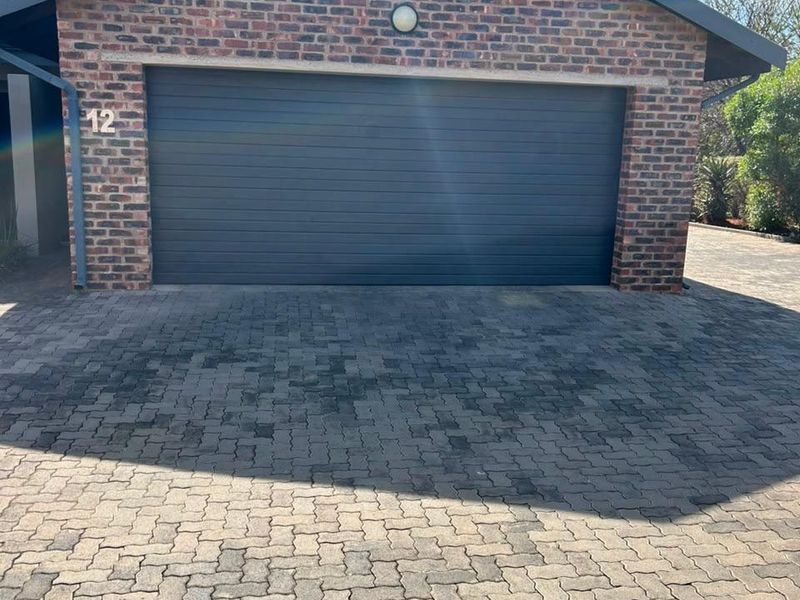 A well-maintained townhouse garage featuring a dark sectional door, exposed brick facade, and a paved driveway area.