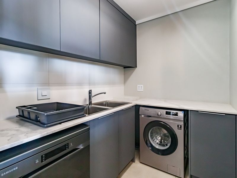 Modern laundry area featuring sleek dark grey cabinetry, stone countertops, a stainless steel sink, and integrated appliance space.