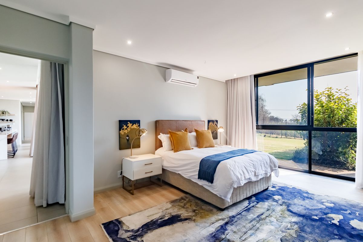 A bright and modern bedroom featuring light timber flooring, recessed lighting, a split-system air conditioner, and large windows with views of the garden.